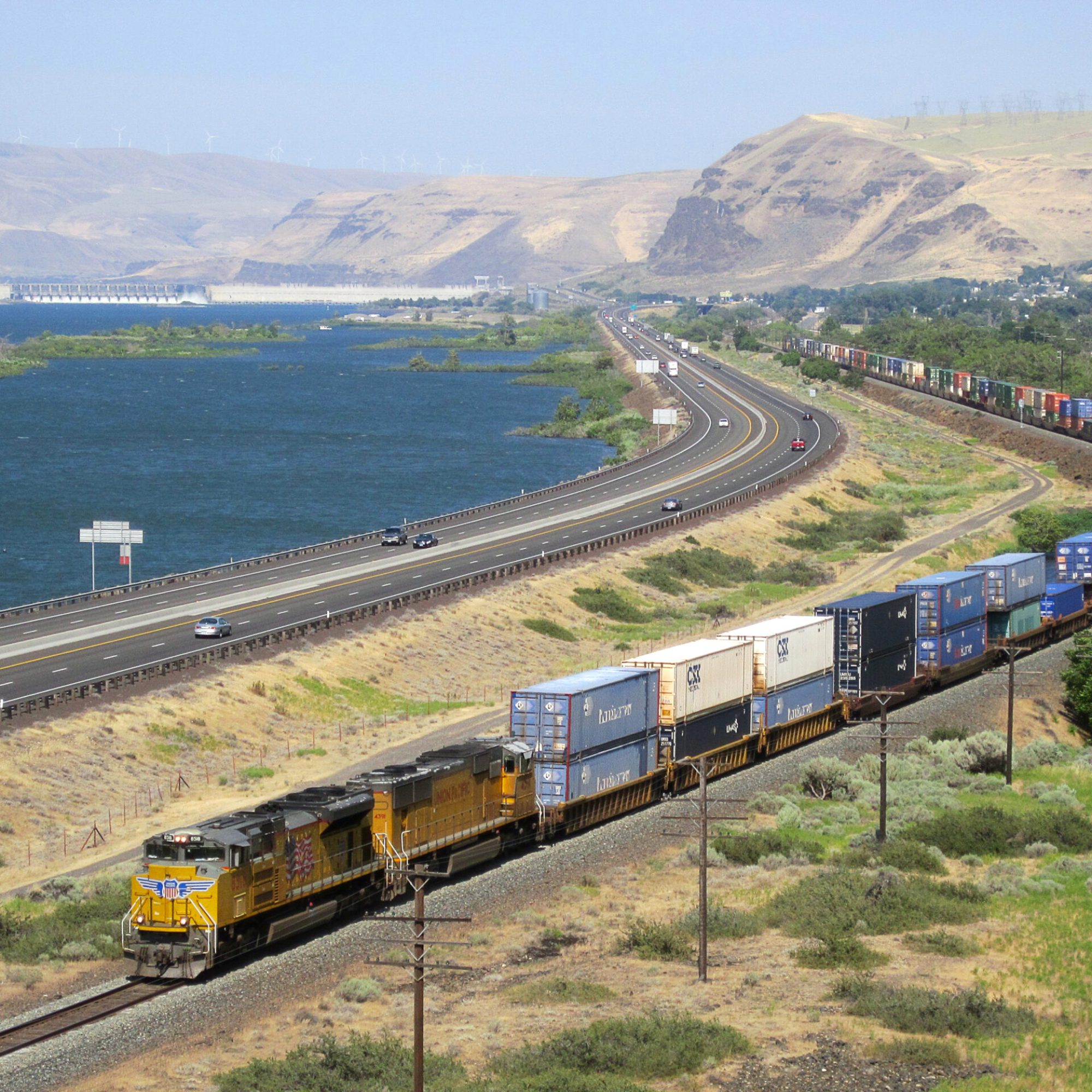 UP 8518 hauls intermodal cars parallel to telephone poles and a major highway. The background features wind turbine topped hills and the Columbia River emerging from the John Day Dam.